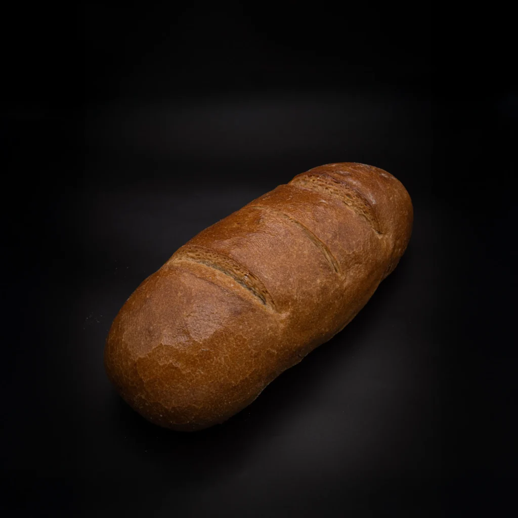 Ein Laib Brot mit glänzender brauner Kruste und drei diagonalen Schlitzen auf der Oberseite auf einem dunklen, festen Hintergrund.