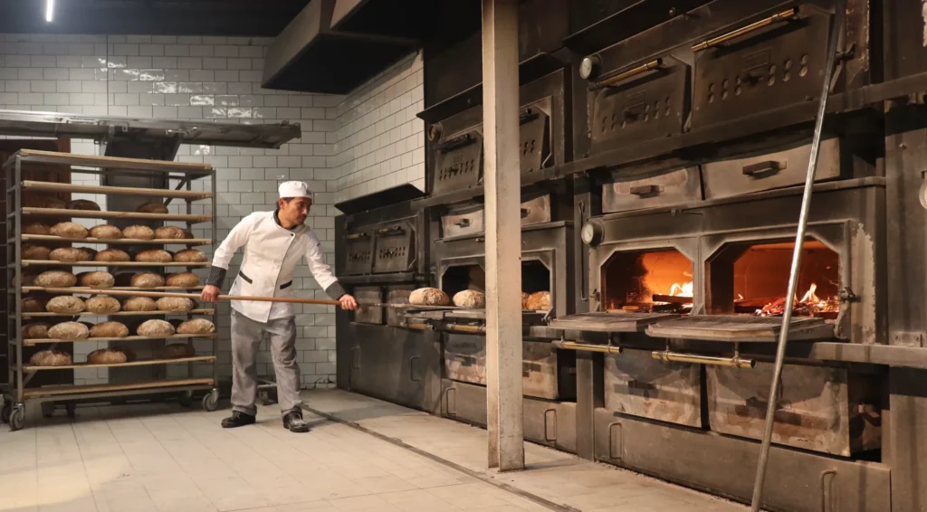 Ein Bäcker in weißer Uniform und mit Hut schiebt mit einer langen hölzernen Schaufel Brotlaibe in einen großen, traditionellen Steinofen mit sichtbaren Flammen. Neben ihm stehen in einer gekachelten Backstube Regale voller Brot.