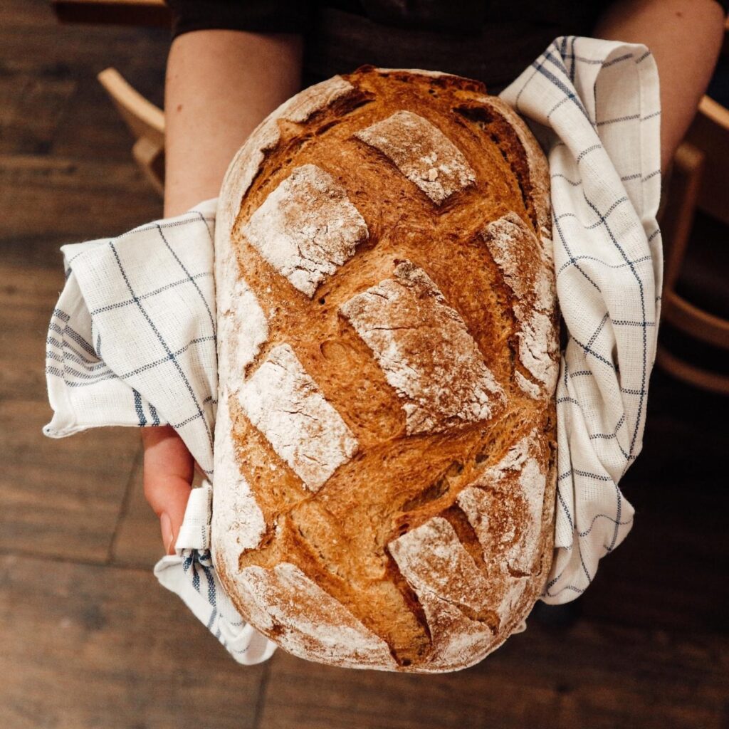 Eine Person hält einen frisch gebackenen, rustikalen Laib Brot auf einem weiß-blau karierten Tuch, im Hintergrund ein Holzboden.