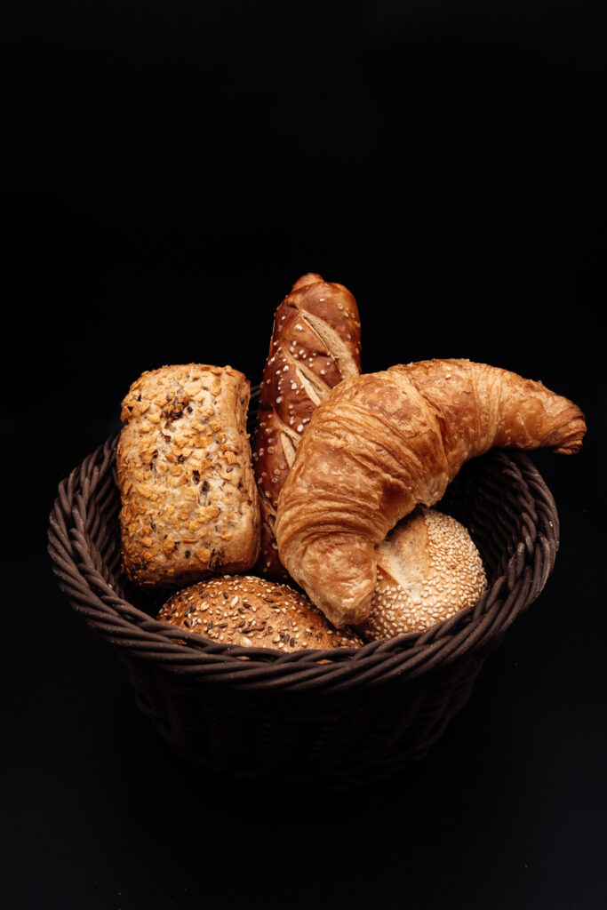 Ein geflochtener Korb, gefüllt mit verschiedenen Brotsorten, darunter ein Croissant, Mehrkornbrötchen und ein Sesambrötchen, vor einem schwarzen Hintergrund.