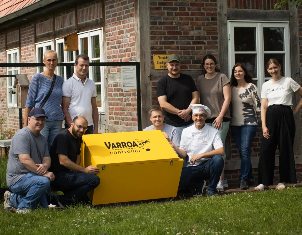 Eine Gruppe von zehn Personen posiert im Freien vor einem Backsteingebäude mit einem großen, leuchtend gelben Kasten mit der Aufschrift Varroa Controller. Einige stehen, andere knien neben dem Kasten, alle lächeln in die Kamera.