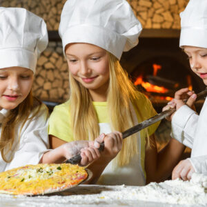 Drei junge Mädchen in Kochmützen und Uniformen backen gemeinsam Pizza. Ein Mädchen in einem gelben Hemd hilft einem anderen, eine Pizza auf ein Holzpaddel zu legen, während das dritte Mädchen Mehl streut. Im Hintergrund leuchtet ein holzbefeuerter Ofen.