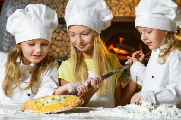 Drei junge Mädchen in Kochmützen und Uniformen backen gemeinsam Pizza. Ein Mädchen in einem gelben Hemd hilft einem anderen, eine Pizza auf ein Holzpaddel zu legen, während das dritte Mädchen Mehl streut. Im Hintergrund leuchtet ein holzbefeuerter Ofen.