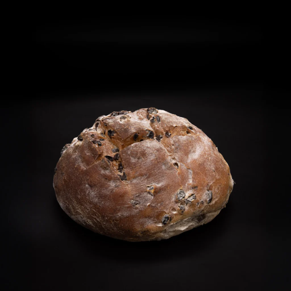Ein runder, rustikaler Brotlaib mit goldbrauner Kruste und sichtbaren Rosinen oder getrockneten Früchten, der auf einem dunklen, schwarzen Untergrund ruht.
