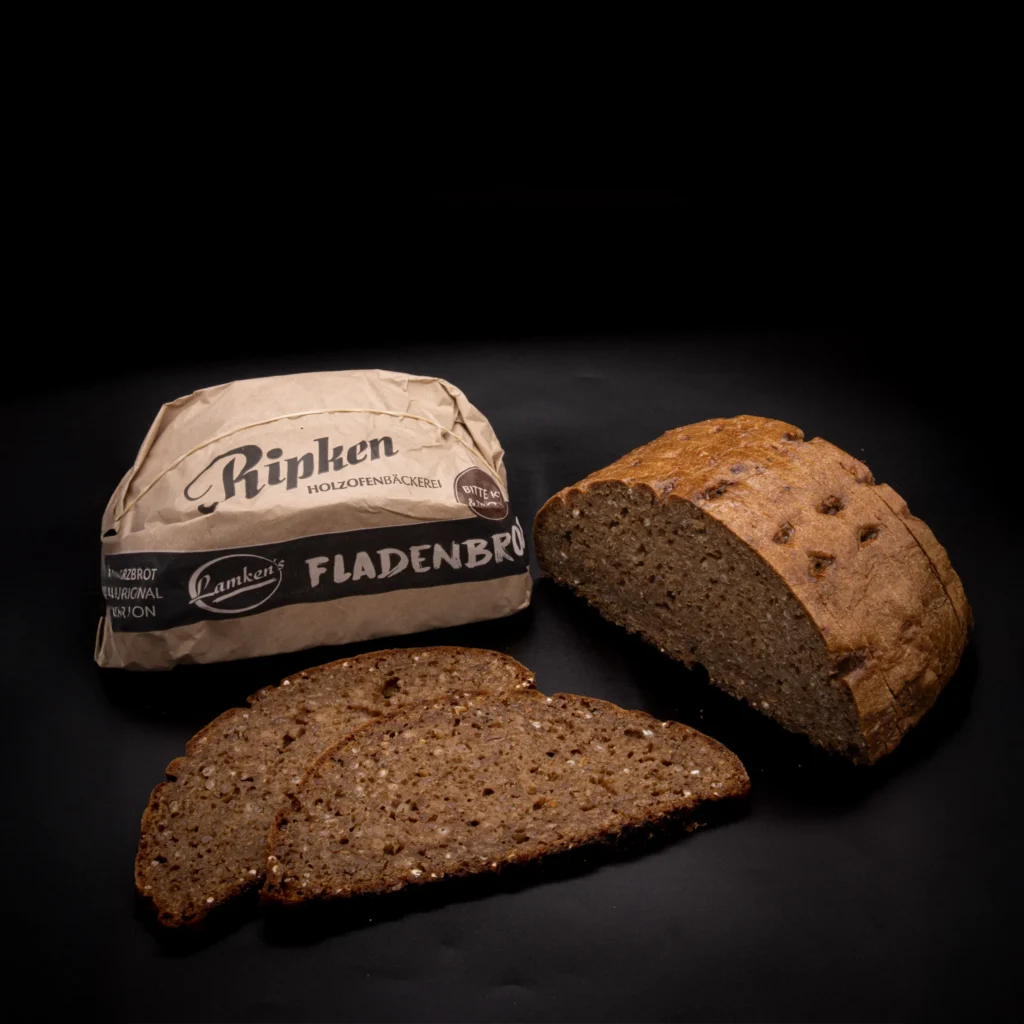 Ein Laib braunen Saatenbrots, teilweise aufgeschnitten, mit drei Scheiben davor. Die Brotverpackung mit den Aufschriften "Ripken" und "Fladenbrot" liegt neben dem Brot auf schwarzem Hintergrund.