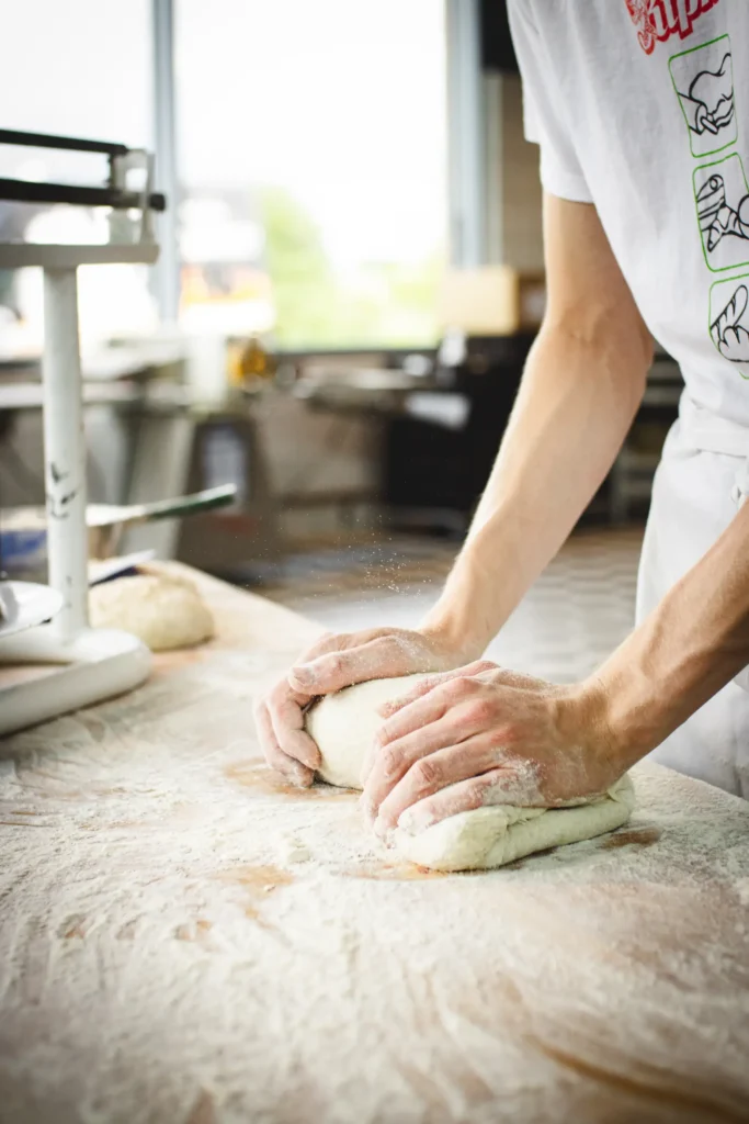 Eine Person knetet in einer hellen Küche mit weißem Hemd und Schürze Teig auf einer bemehlten Fläche. Auf der Arbeitsplatte sind eine Waage und weitere Teigstücke zu sehen.