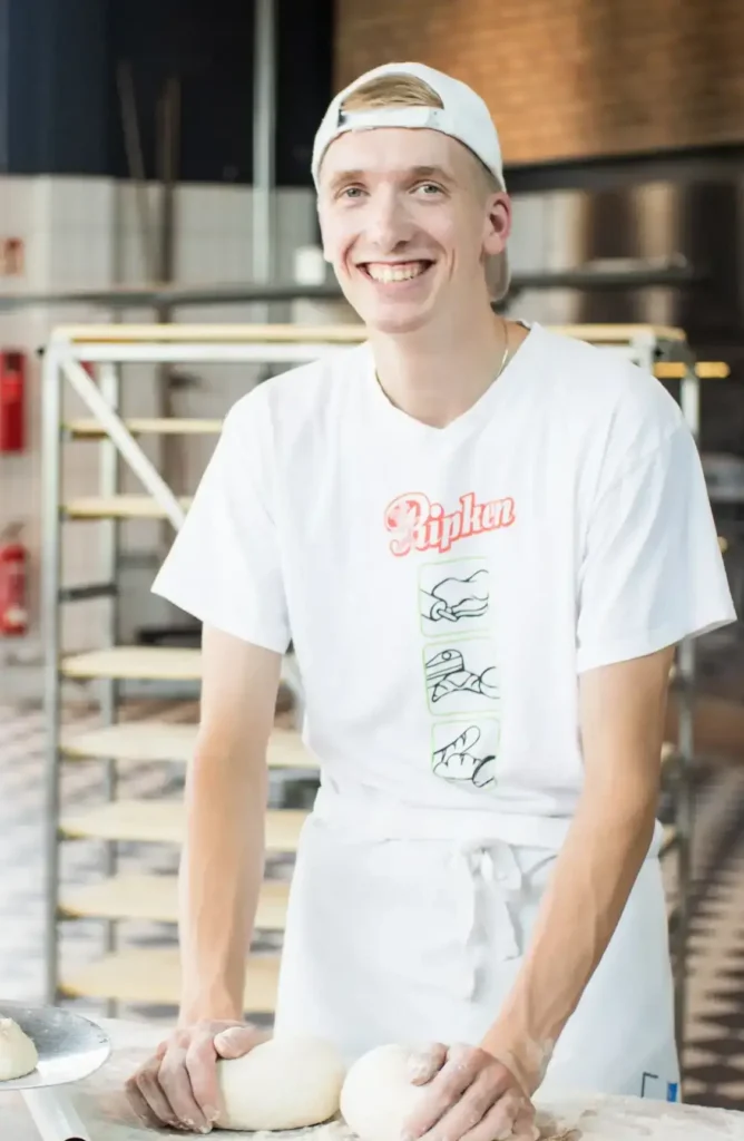 Ein lächelnder junger Mann in einem weißen T-Shirt und einer Mütze steht an einem Tisch und formt Teigkugeln in einer hellen Bäckerei mit Metallgestellen im Hintergrund.