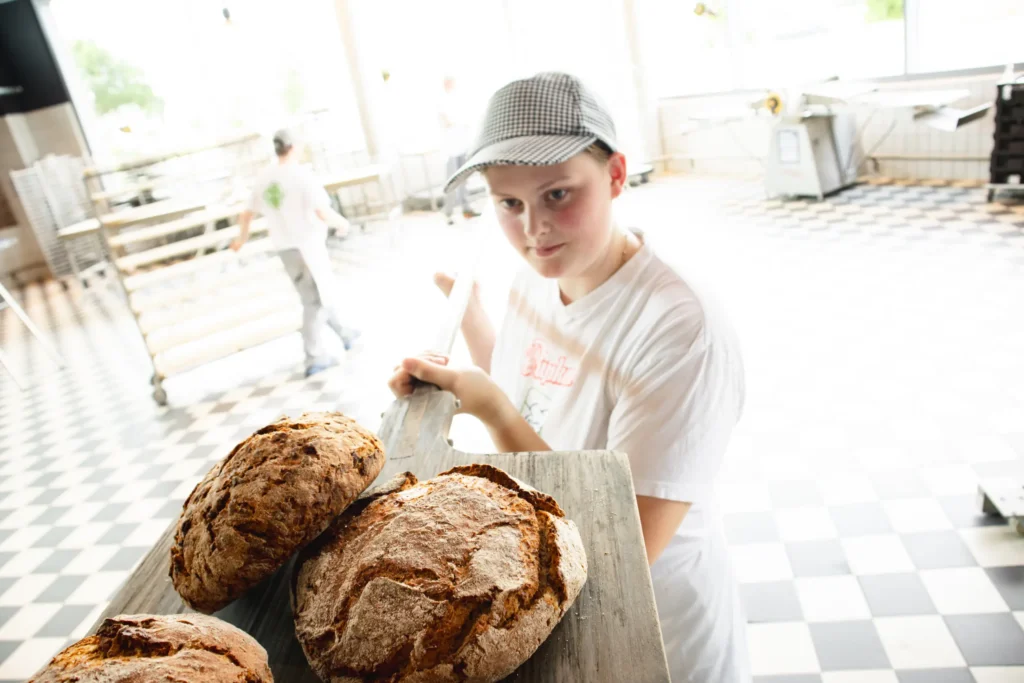 Ein junger Bäcker mit weißem Hemd und karierter Mütze schiebt in einer hellen, gekachelten Bäckereiküche zwei große Laibe rustikalen Brotes mit einem Holzschäler hin und her.