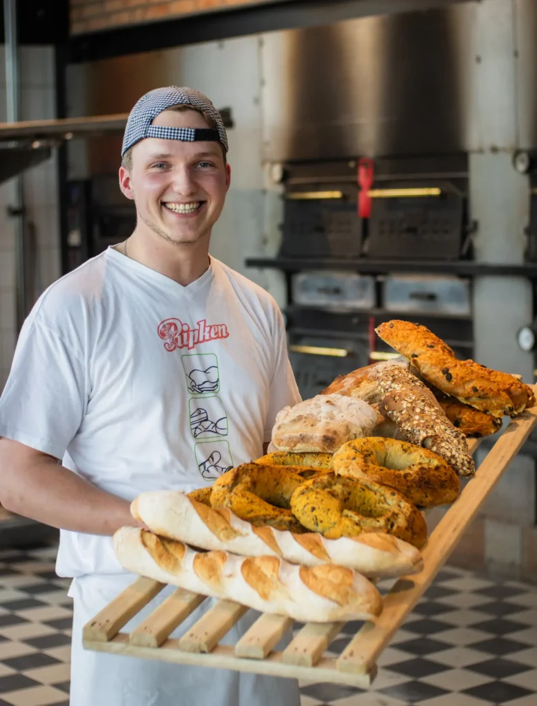 Ein lächelnder Bäcker in einem weißen T-Shirt und einer Mütze hält ein großes Holztablett mit verschiedenen frisch gebackenen Broten in der Bäckerei Ripken mit Öfen im Hintergrund.
