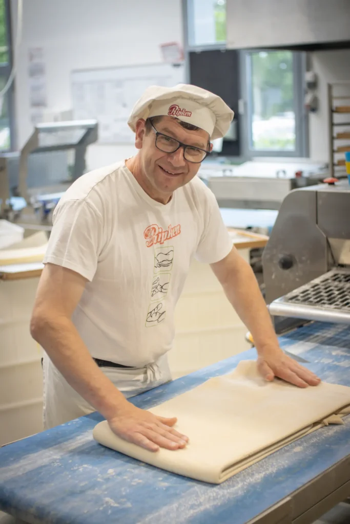 Ein lächelnder Bäcker, der eine Brille, einen weißen Hut und ein weißes T-Shirt trägt, formt Teig auf einer blau bemehlten Arbeitsfläche in einer hellen Bäckereiküche. Im Hintergrund sind Bäckereigeräte und Fenster zu sehen.