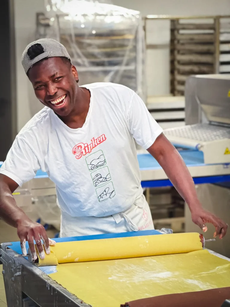 Ein Mann mit weißem T-Shirt und Mütze lächelt, während er auf einem Arbeitstisch in einer Großküche gelben Teig ausrollt. Im Hintergrund sind Backgeräte und Backbleche zu sehen.