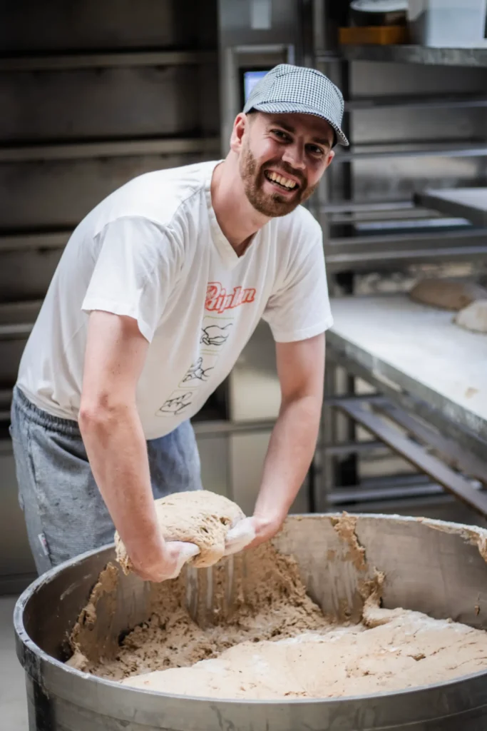 Ein Mann, der eine Mütze, ein T-Shirt und eine Jogginghose trägt, lächelt, während er in einer großen Metallschüssel in der Bäckerei Ripken Sauerteig ansetzt.