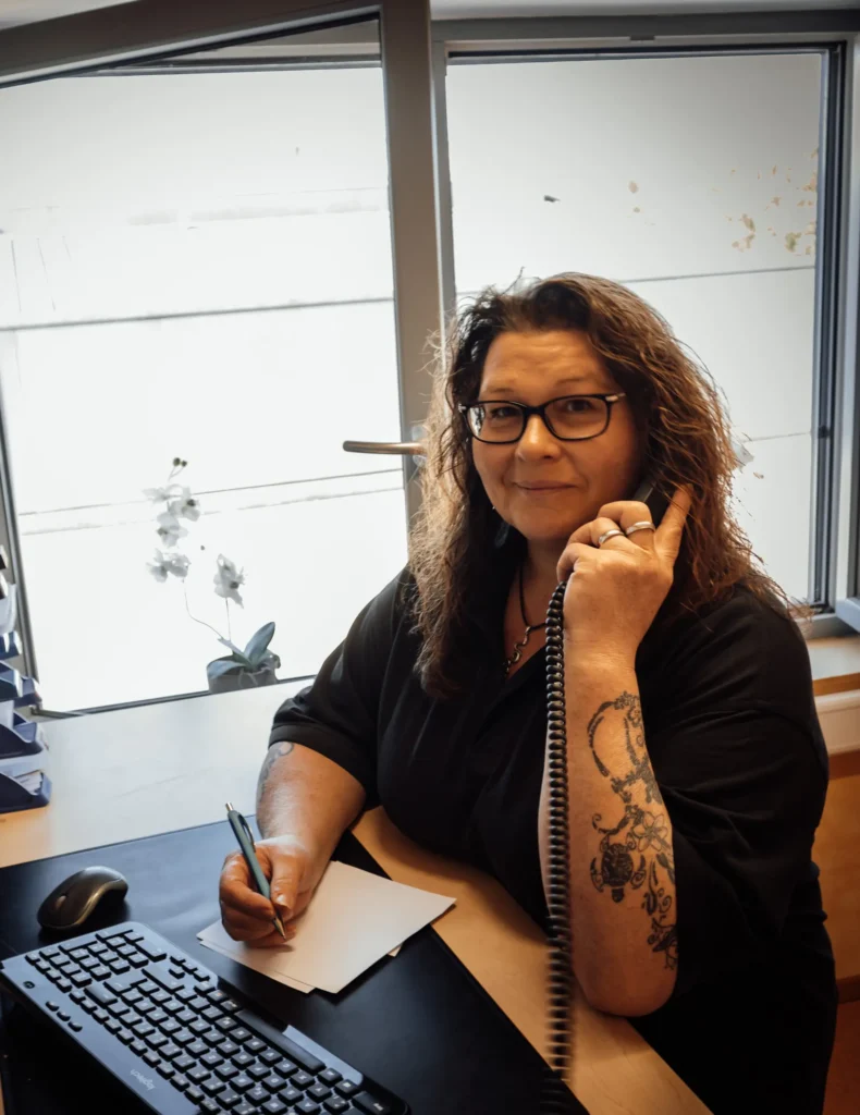 Eine Frau mit lockigem Haar und Brille sitzt an einem Schreibtisch im Büro der Bäckerei Ripken, hält ein Telefon an ihr Ohr und schreibt auf einem Notizblock. Vor ihr befindet sich eine Tastatur, und im Hintergrund ist ein Fenster mit einer Topforchidee zu sehen.