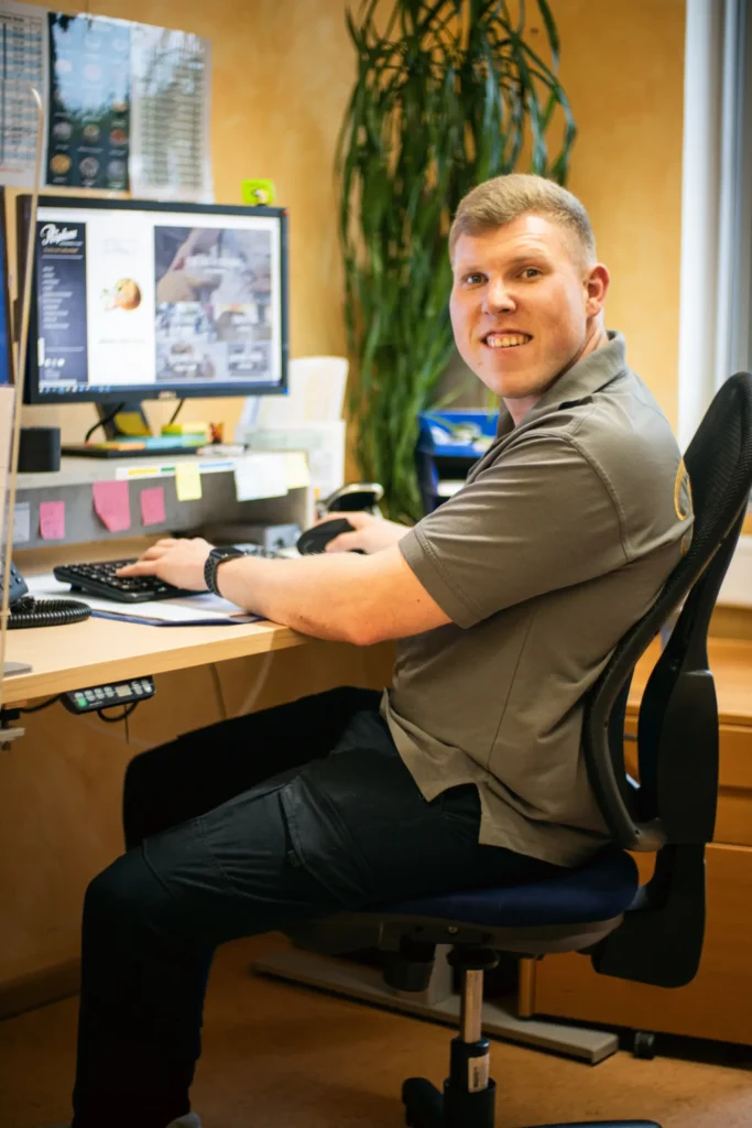 Ein Mann sitzt an einem Schreibtisch im Büro der Bäckerei Ripken, tippt auf einer Tastatur und lächelt in die Kamera. Er trägt ein graues Poloshirt und eine dunkle Hose. Im Hintergrund sind ein Computer, Büromaterial und eine große Pflanze zu sehen.