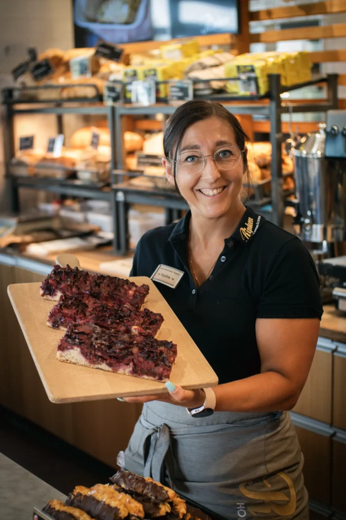 Eine lächelnde Frau mit Brille und schwarzem Poloshirt hält ein Holztablett mit Kuchen in der Bäckerei Ripken, im Hintergrund stehen Regale mit Brot.