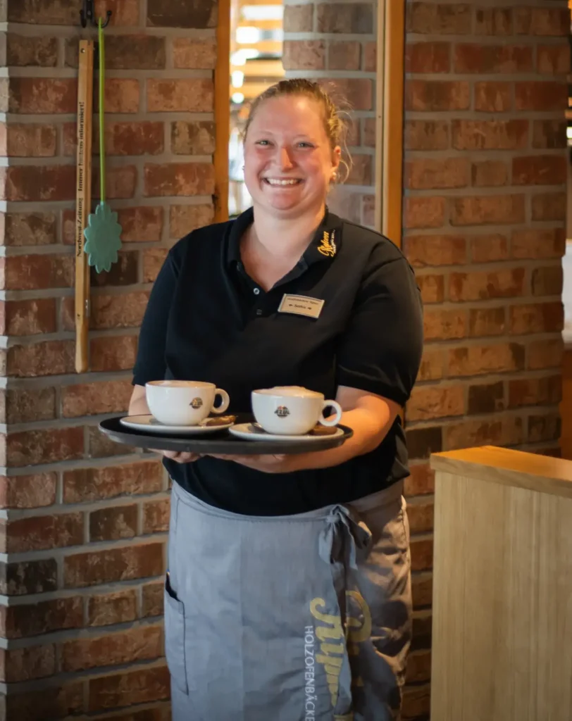 Eine lächelnde Frau in dunklem Hemd und grauer Schürze hält ein Tablett mit zwei Tassen Kaffee in einem gemütlichen Café mit Backsteinwänden.