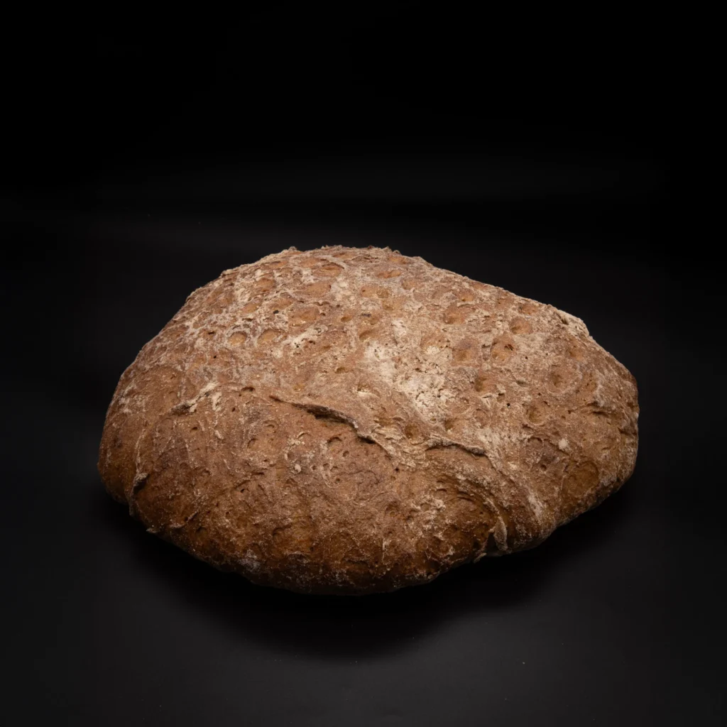 Ein runder Laib rustikales Schwarzbrot liegt auf einer schwarzen Fläche mit dunklem Hintergrund, der seine strukturierte Kruste hervorhebt.
