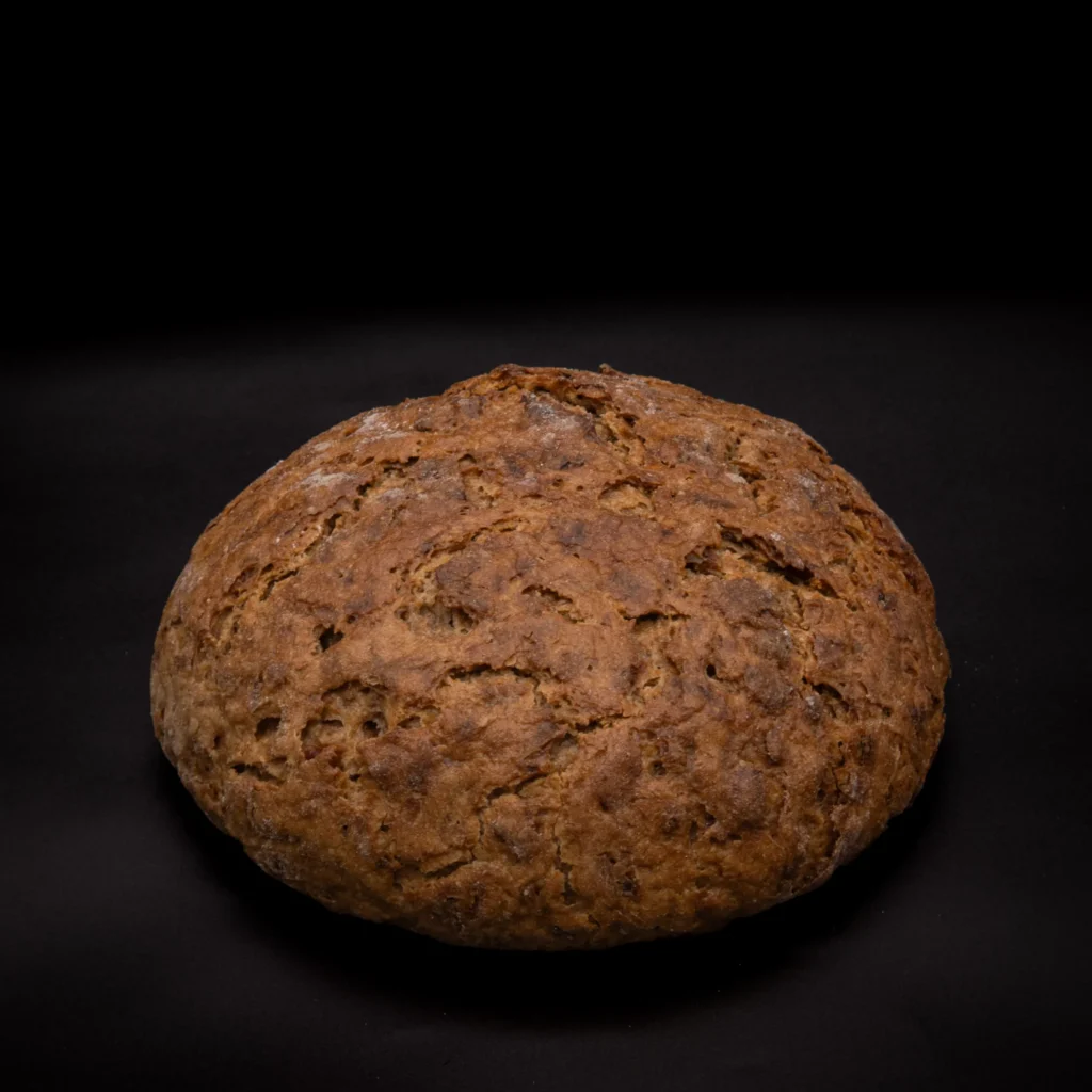 Ein runder Laib rustikales Schwarzbrot liegt auf einer dunklen Fläche mit schwarzem Hintergrund, der seine raue, strukturierte Kruste hervorhebt.