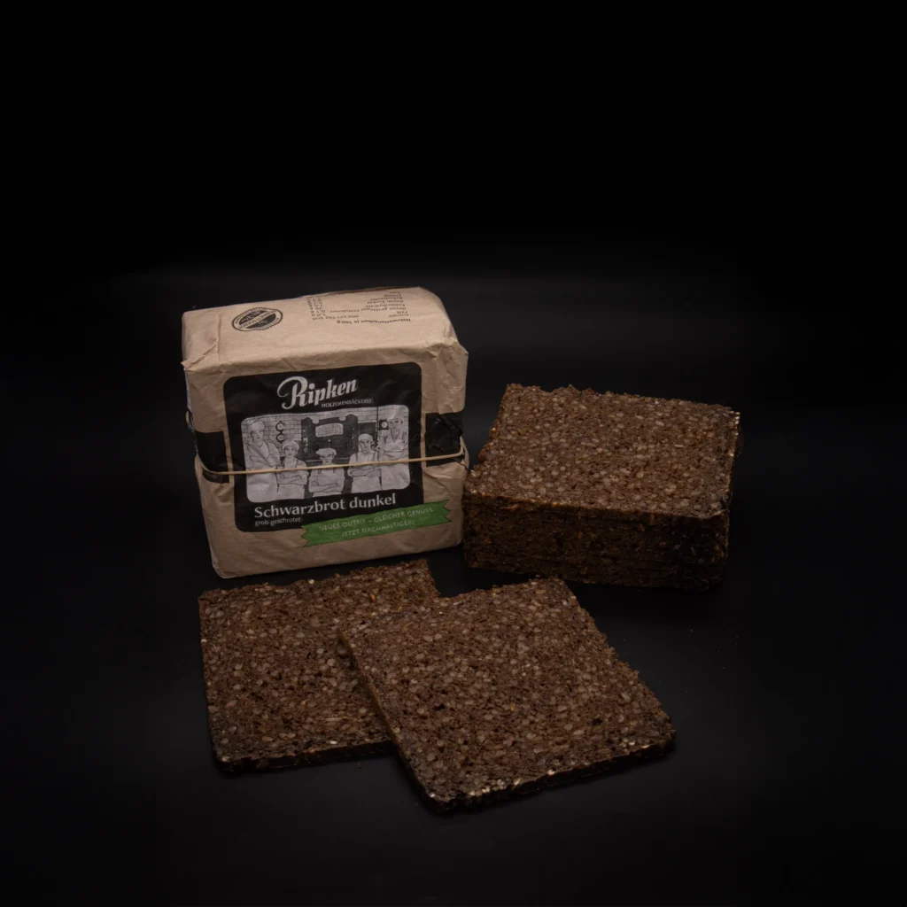 Auf einer schwarzen Fläche liegt eine Packung Ripken Schwarzbrot dunkel. Davor liegen drei große, quadratische Scheiben des dichten, groben Brotes.