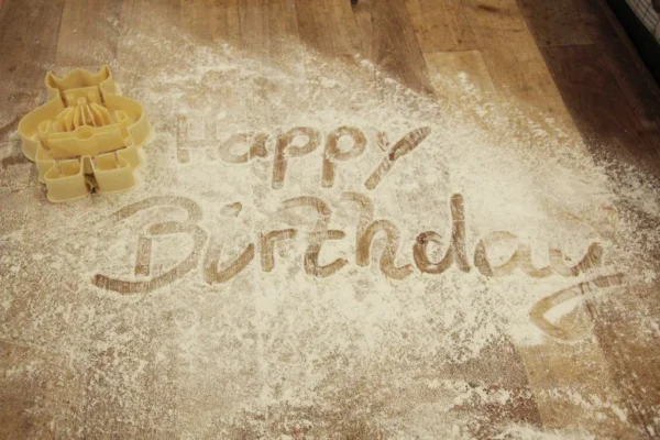 Happy Birthday" wird mit Mehl auf eine hölzerne Fläche geschrieben, und in der Nähe befinden sich Ausstechformen in Form eines Geburtstagskuchens.