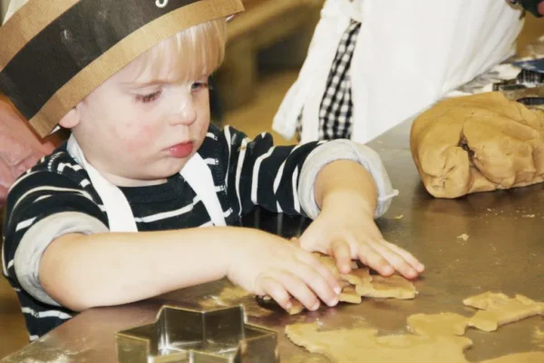 Ein kleines Kind mit gestreiftem Hemd und Papierkochmütze drückt mit beiden Händen Plätzchenteig aus und formt ihn auf einem Tisch. Ausstechformen und weiterer Teig liegen in der Nähe.