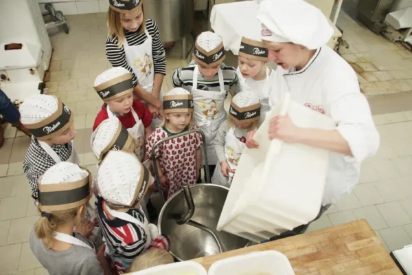 Eine Gruppe von Kindern mit Kochmützen und Schürzen versammelt sich um einen erwachsenen Koch, der in einer Küche eine Zutat in eine große Metallschüssel schüttet.