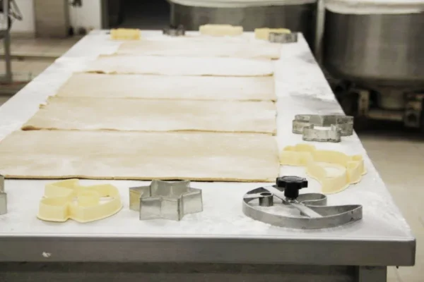 Ausgerollte Teigblätter auf einem bemehlten Metalltisch mit verschiedenen Ausstechern und Backwerkzeugen, die um die Ränder herum angeordnet sind, in einer Industrieküche oder Bäckerei.