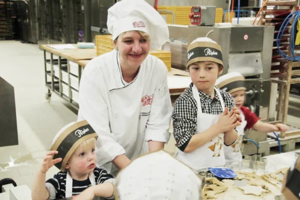 Ein lächelnder Bäcker mit einer weißen Kochmütze hilft drei kleinen Kindern, die ebenfalls Papiermützen tragen, an einem Tisch in einer Bäckerei Teig zu formen. Die Kinder sind mit der Arbeit beschäftigt, und auf dem Tisch sind Backutensilien zu sehen.