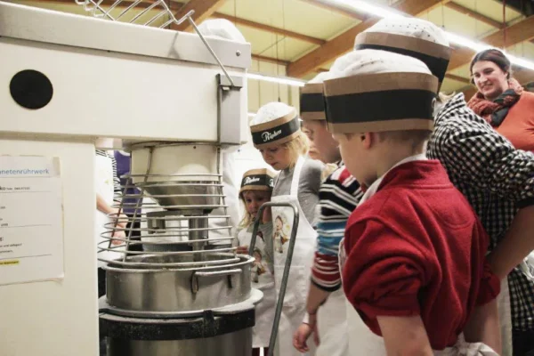 Mehrere Kinder mit Schürzen und Papierhüten beobachten einen großen Industriemixer in einer Bäckerei, beaufsichtigt von einer erwachsenen Frau mit Schal und kariertem Hemd. Die Gruppe scheint mit einer Backtätigkeit beschäftigt zu sein.