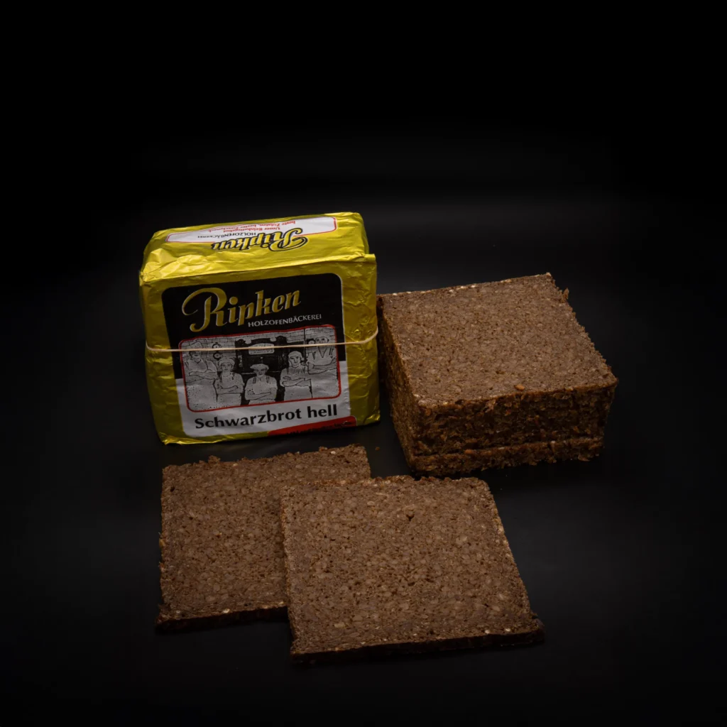 Eine Packung Rinken Schwarzbrot hell ist neben mehreren quadratischen Scheiben des dichten, dunklen Brotes auf schwarzem Hintergrund abgebildet.