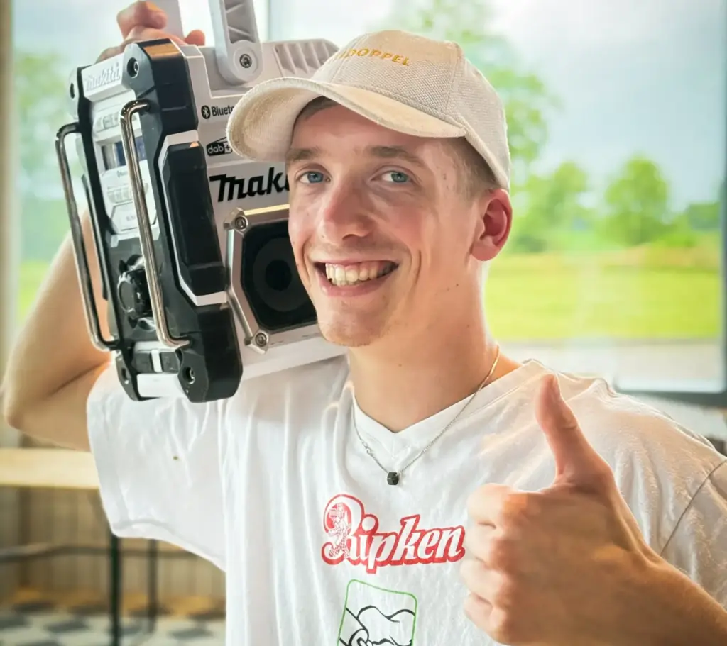Ein lächelnder junger Mann mit weißer Mütze und T-Shirt, der ein großes Kofferradio auf der Schulter trägt und den Daumen nach oben streckt, steht in einem Innenraum und blickt unscharf auf die grüne Landschaft im Hintergrund.