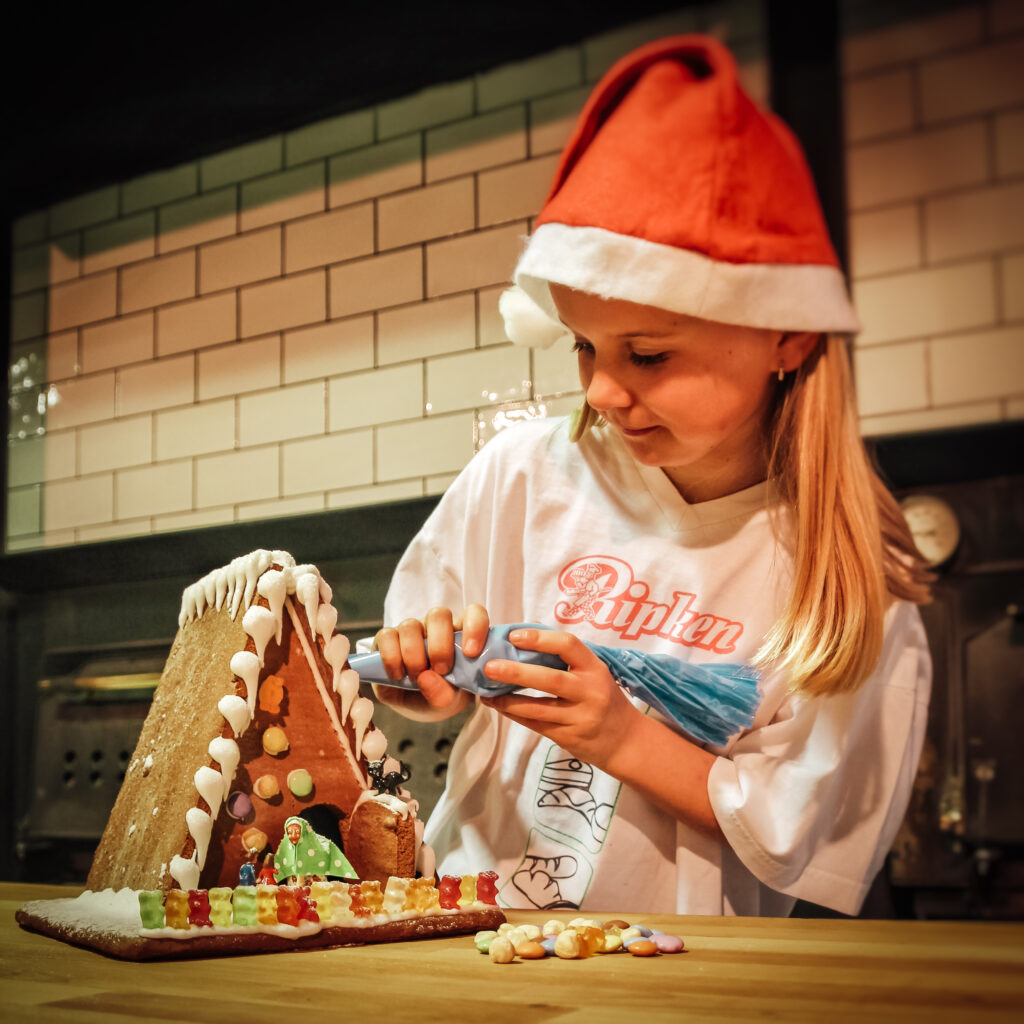Ein junges Mädchen mit einer Weihnachtsmannmütze dekoriert ein Lebkuchenhaus mit Zuckerguss und bunten Süßigkeiten auf einem Küchentisch, lächelnd und konzentriert auf ihre Arbeit.