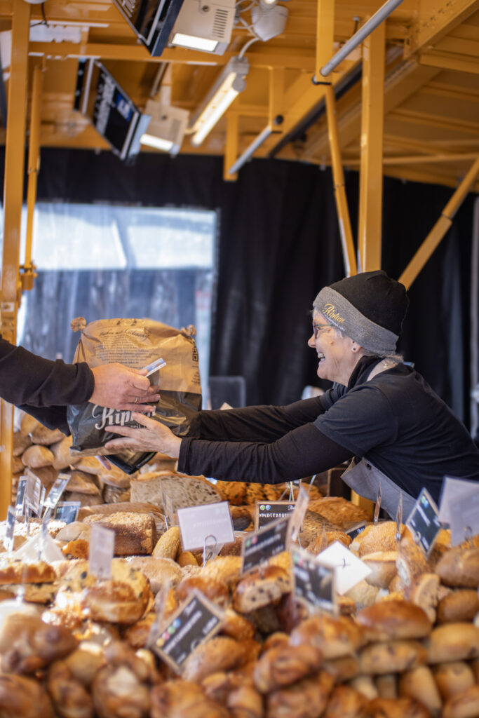 Ein lächelnder Verkäufer mit Mütze reicht einem Kunden an einem Marktstand im Freien, der mit verschiedenen Brotsorten und Gebäck gefüllt ist, eine Tüte mit Backwaren.