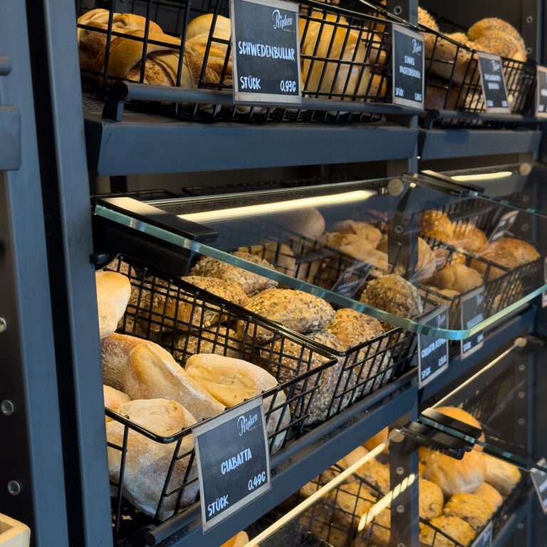 In einer Bäckerei sind die Regale mit verschiedenen Sorten frischer Brötchen und Brote in schwarzen Drahtkörben gefüllt, jede Abteilung ist mit einem Schild versehen, auf dem die Namen und Preise der Brotsorten stehen.
