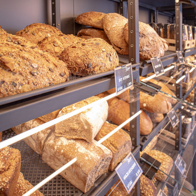 In einer Bäckerei stehen Regale mit verschiedenen Brotsorten, darunter auch kernige und knusprige Sorten. Jedes Regal ist mit Preisschildern versehen, die vor den verschiedenen Brotsorten angebracht sind.