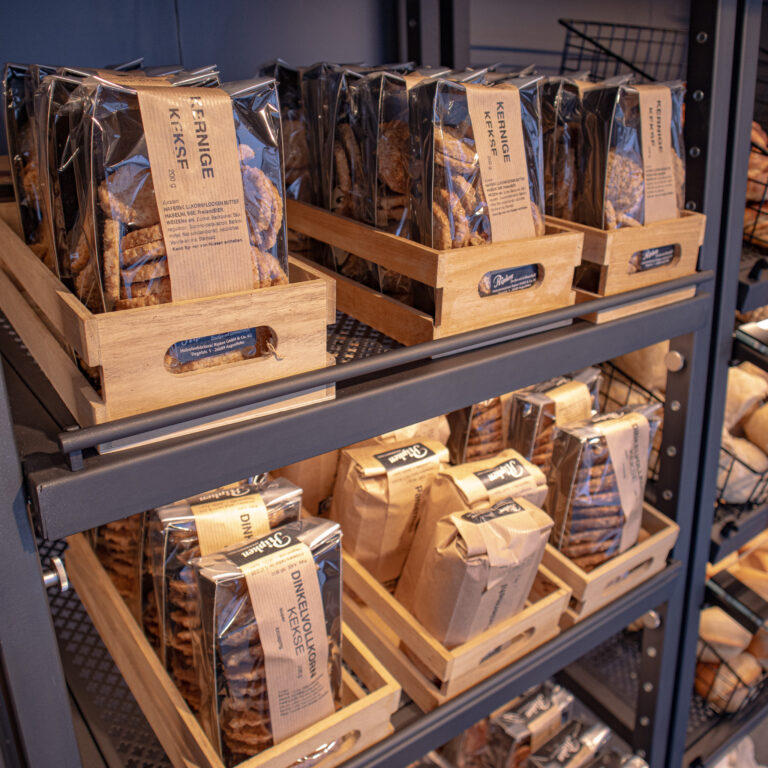 Holzkisten, gefüllt mit verpackten Keksen, stehen fein säuberlich auf metallenen Bäckereiregalen. Jede Packung trägt ein braunes Etikett mit deutschem Text. Weitere Backwaren sind in Körben in der Nähe zu sehen.