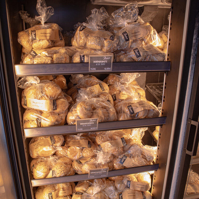 In einer Kühlvitrine stapeln sich die Brötchentüten auf Metallregalen. Jede Tüte ist etikettiert, und die Regale sind mit Preisschildern versehen. Die Brötchen sehen frisch aus und sind in durchsichtigen Plastikbeuteln verpackt.