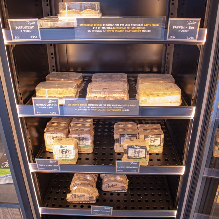 Eine Bäckereivitrine mit verpacktem Gebäck und Kuchen in Regalen, darunter Artikel mit der Aufschrift "Portugieser" und "Kuchen-Box". An jedem Regal sind Preis- und Produktschilder zu sehen.