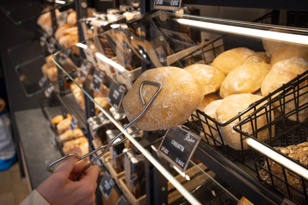 Eine Person nimmt mit einer Zange ein rundes Ciabatta-Brötchen aus einer Bäckereivitrine, die mit verschiedenen frischen Brötchen gefüllt ist, unter hellem Licht.