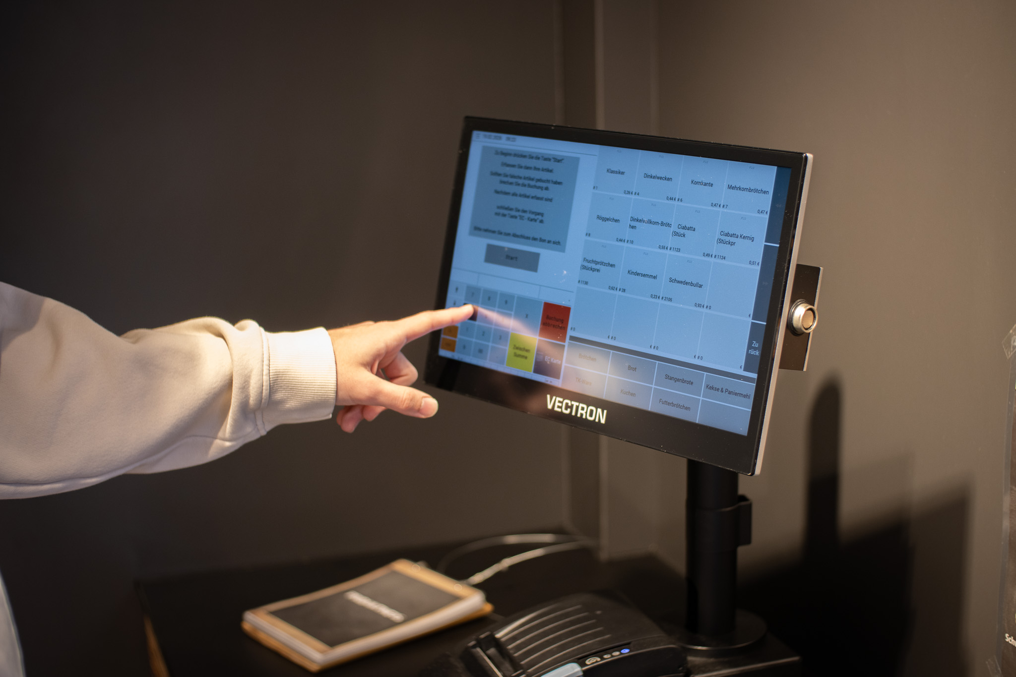 Eine Person, die einen hellen Ärmel trägt, berührt den Bildschirm eines Vectron-Kassenterminals. Ein kleines Gerät und ein Notizblock befinden sich auf dem Tresen unter dem Bildschirm.