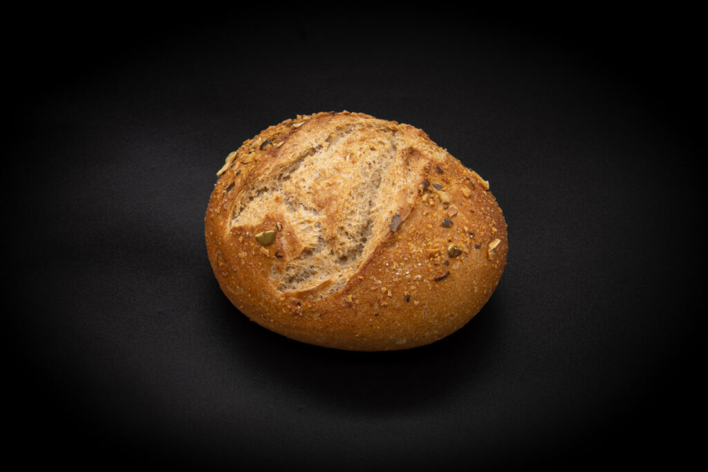 Auf einem schwarzen Hintergrund liegt ein rundes Brötchen mit goldbrauner Kruste und Körnern darauf.