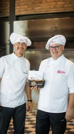 Zwei lächelnde Bäcker in weißen Uniformen und Hüten stehen in einem Haus; einer hält eine Tüte mit der Aufschrift Pinken. Sie stehen vor einem gemauerten Ofen und einem karierten Boden, der an eine Bäckerei denken lässt.