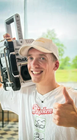 Ein lächelnder junger Mann mit weißer Mütze und T-Shirt hält einen tragbaren Lautsprecher auf seiner Schulter und zeigt den Daumen nach oben. Er befindet sich in einem Innenraum mit großen Fenstern, die eine grüne Landschaft im Hintergrund zeigen.