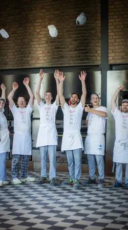 Sechs Bäcker in weißen Hemden und Schürzen stehen in einer Bäckerei, lächeln und heben ihre Arme, während sie ihre Hüte in die Luft werfen. Im Hintergrund sind Backsteinwände und Industrieöfen zu sehen.