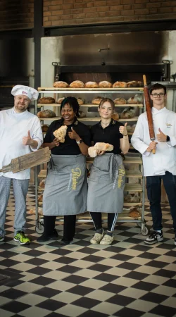 Vier Bäcker stehen lächelnd vor den mit Brot gefüllten Regalen. Zwei Frauen in Schürzen halten die Brote, während zwei Männer in weißen Kochjacken die Backwerkzeuge halten. Alle zeigen den Daumen nach oben auf einem karierten Boden.